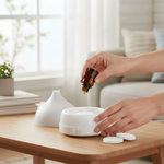 Hands adding oil to a white tabletop diffuser with two round white replacement pads visible on the table