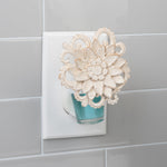 Electric fragrance plug-in inserted in an outlet with a decorative cream floral cover and a visible blue refill bottle.