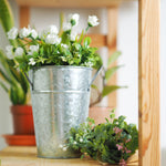 Galvanized metal bucket planter holding white floral arrangement on a wooden shelf with plants in background