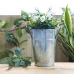 Galvanized metal bucket planter filled with white flowers and greenery on a wooden tabletop with foliage behind