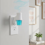 Plug-in fragrance diffuser inserted in a wall outlet with a glass bottle of blue-green oil attached and decorative artwork in the background.