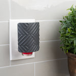 Mainstays textured black fragrance plug inserted in a wall outlet next to a potted plant on a tiled wall.