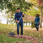 Man raking leaves on a grassy slope using a Fiskars leaf rake while another person carries a leaf bag in background