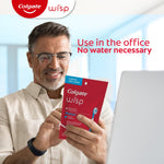 Smiling man holding a red Colgate Wisp pouch while seated at a desk, overlaid text reads 'Use in the office No water necessary'.