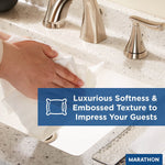 Hand using a white embossed Marathon multifold towel to wipe a sink, blue banner text about softness and texture visible