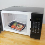 Black lidded meal prep container with salmon and vegetables inside a microwave oven on a wooden counter.