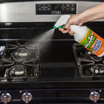 Person spraying Mean Green Super Strength cleaner onto a stovetop and gas burners from a white trigger bottle