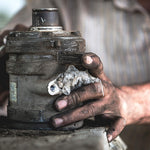 Greasy, dirt-covered hands holding a heavily soiled mechanical component against a blurred background.