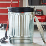 Metal trash can with a black garbage bag liner tied at the top, broom and garage shelving in background