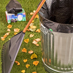 Outdoor lifestyle shot showing a metal trash can lined with a black drawstring bag, a rake and a product box in the grass.