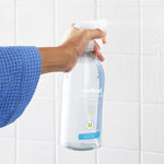 Hand holding a clear Method spray bottle in front of tiled shower wall, demonstrating product in use.