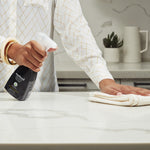 Lifestyle photo of a person spraying a black Method Granite Polish bottle onto a light marble countertop while wiping with a cloth.