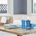 Lifestyle scene on a wooden table with folded towels, a wicker basket and Method laundry products including a blue detergent bottle.