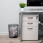 Black metal mesh trash can placed next to a white office desk filled with crumpled paper
