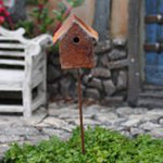Miniature rust-colored birdhouse on a stake placed in a landscaped scene with stonework and a bench in background
