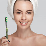 Smiling woman holding a green-handled toothbrush beside her face, hair wrapped in a white towel