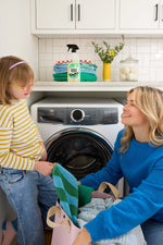 Laundry room scene with Molly's Suds spray bottle on a shelf while an adult and child handle towels by a front-load washer