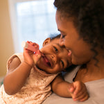 Smiling mother holding a laughing baby who is playfully holding a pink toothbrush against the mother's cheek.