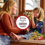 Warm lifestyle photo of an adult and child on a windowsill sharing and smelling garden flowers with a circular garden-scents message.