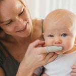 Mother smiling while gently using a silicone finger toothbrush on an infant, close-up showing toothbrush texture and baby’s face.