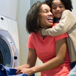 Smiling adult and child hugging near a washing machine in a laundry room, lifestyle image without visible packaging text.
