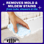 Hand using a blue Mr. Clean Magic Eraser pad to clean tub or shower grout area with suds and drain visible