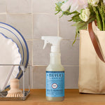 Styled kitchen scene showing a Mrs. Meyer's Rain Water spray bottle between dishes and a tote with flowers.