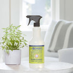 Lifestyle photo of Mrs. Meyer's Clean Day glass cleaner bottle on a small table with a potted plant and blurred window background