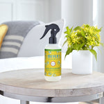 Lifestyle photo of Mrs. Meyer’s honeysuckle room freshener bottle on a wooden table beside a potted green plant