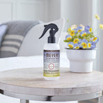 Mrs. Meyer’s Clean Day Compassion Flower room freshener bottle staged on a table next to a small pot of pansy-like flowers