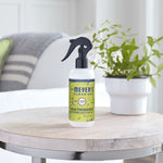 Mrs. Meyer’s Lemon Verbena room freshener placed on a wooden table beside a potted plant in a bright room setting.