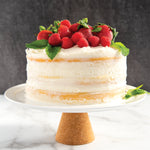 Rustic naked-style layer cake with white frosting and fresh raspberries and mint leaves on a white cake plate.