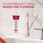 Native cinnamon & mint toothpaste tube standing on a sink counter beside two toothbrushes in a glass with tiled wall background