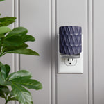 Navy textured ScentPlug diffuser inserted into a wall outlet on beadboard wall with a plant at left