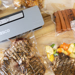 Top-down view of a NESCO vacuum sealer and multiple vacuum-sealed bags holding seasoned steaks, vegetables, and sausages on a wooden surface.