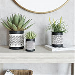 Three black-and-white patterned ceramic planters styled on a wooden console with faux succulents and a round wall mirror.