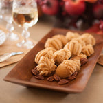 Wooden tray of small autumn-shaped cakelets and pecans with blurred glassware and fruit in background