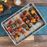 Aluminum roasting pan with black rack holding skewered beef and vegetable kebabs on a wooden table setting