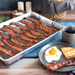 Aluminum roasting pan with nonstick rack holding strips of cooked bacon arranged on a styled tabletop