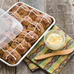 Quarter sheet pan filled with baked yeast rolls drizzled with icing and covered partially by a clear plastic lid, styled with butter on a napkin.