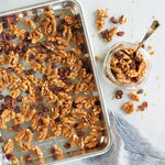 Quarter sheet aluminum pan filled with roasted walnuts and dried fruit on a light surface with a jar of nuts and a striped cloth nearby.
