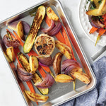 Aluminum quarter sheet pan holding roasted vegetables including zucchini, red onion, carrots and garlic on a light countertop.