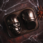 Nordic Ware two-cavity skull cake pan photographed on a dark textured surface with faux spiderwebs and a decorative spider.