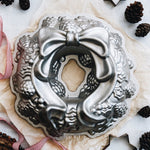 Nordic Ware silver holiday wreath bundt pan shown on parchment with pine cones and ribbon props