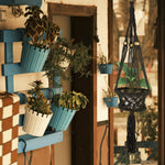 Lifestyle image of a black macramé hanging planter displayed near a rustic porch wall with potted decor