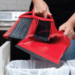 Hands holding red O-Cedar broom and dustpan over a trash bin, demonstrating sweeping into the pan