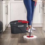 Person mopping a kitchen floor with O-Cedar triangular red mop head and matching black and red EasyWring bucket visible