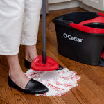Person using an O-Cedar spin mop near a black and red O-Cedar bucket, feet visible on a hardwood floor
