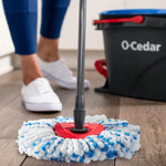 Low-angle close shot of O-Cedar mop head on floor with bucket and person’s lower legs in background