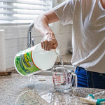 Person pouring OdoBan disinfectant from a gallon jug into a glass measuring cup on a kitchen countertop.